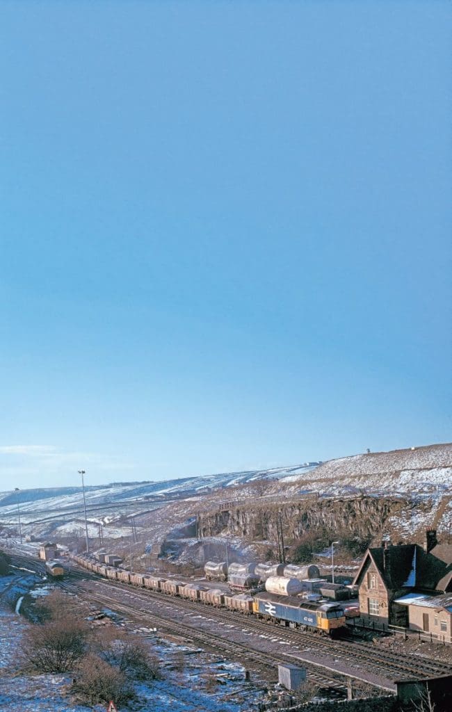  In BR large logo livery, No. 47441 (originally 1557) is seen with a train of two-axle aggregate wagons at Peak Forest on March 2, 1990. Note the two-axle cement wagons in the background and the Class 37 light engine on the left.